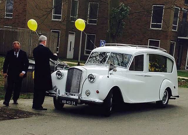 Witte uitvaartauto met gele ballonnen